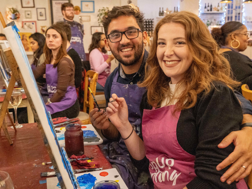 Valentine's Date Ideas - sip and paint. Two people at a sip and paint event, smiling at the camera. There are other guests in the background